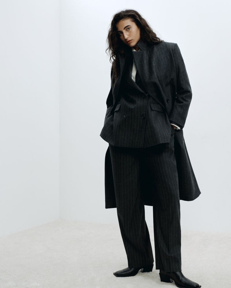 Woman in pinstripe suit poses in white room. She wears a double-breasted blazer, matching trousers, and black heeled boots in studio shot.