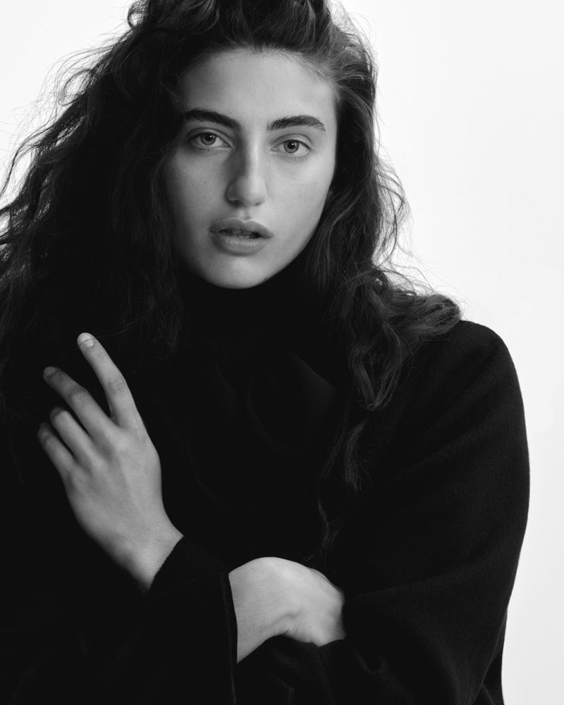 Black and white portrait of a woman with wavy dark hair wearing a turtleneck sweater, in a studio with a neutral background.