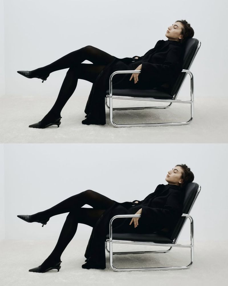 A woman in black attire is elegantly reclined in a chair against a white backdrop, showcasing a blend of fashion and minimalist style.