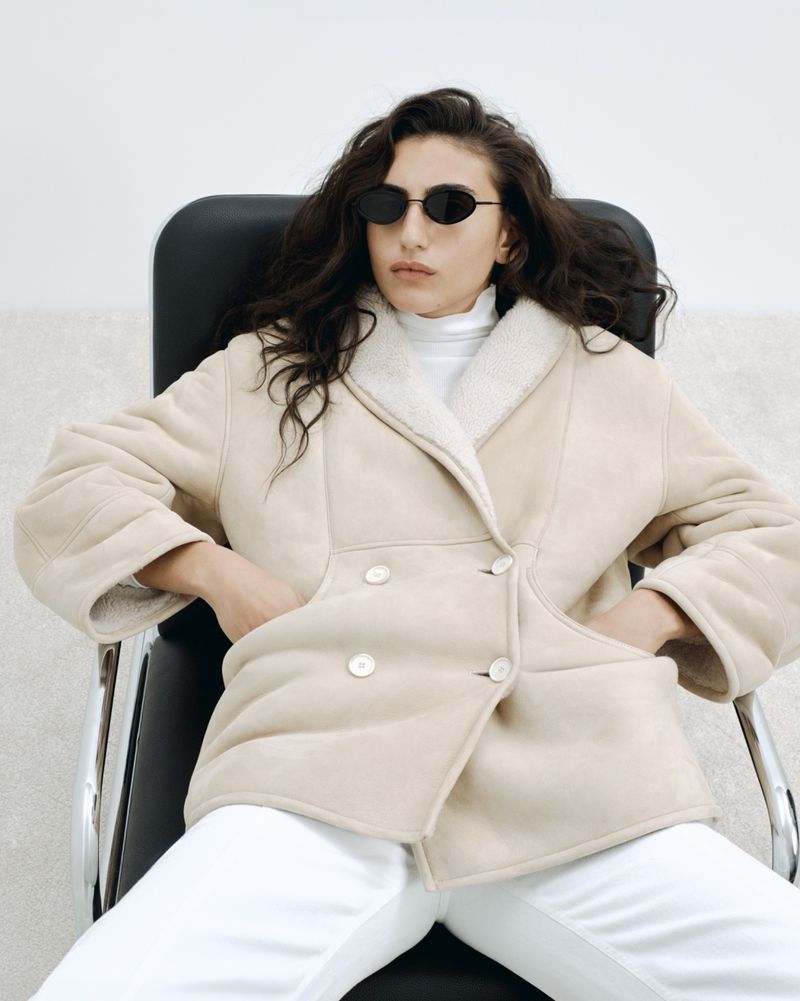 A woman with long dark hair sits in a black chair wearing sunglasses, a light-beige jacket with white pants in a clean, bright indoor setting.