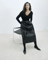 Model Paola Manes seated on a chrome chair wearing a black knit top, voluminous black leather skirt, and pointed heels in a white studio.