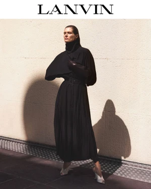 Malgosia Bela posing in a black high-neck pleated belted dress and mule heels against a sunlit wall for Lanvin.