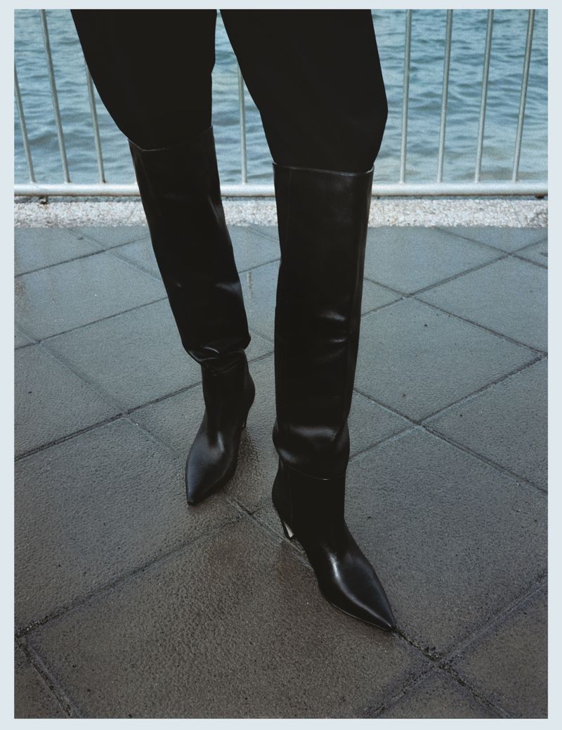 High-angle view of a person wearing black pants and shiny black knee-high pointed boots standing on a gray tiled surface near a railing and water.