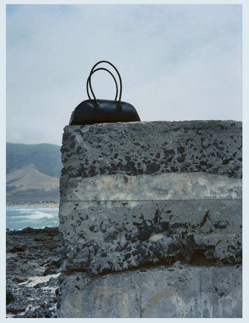 Black leather handbag rests on a weathered concrete block by the sea, with distant mountains and a cloudy sky creating a scenic backdrop.