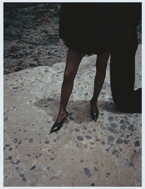 Fashion shot of a person in black tights and shoes, partially visible, standing on a textured stone surface with a rocky background.