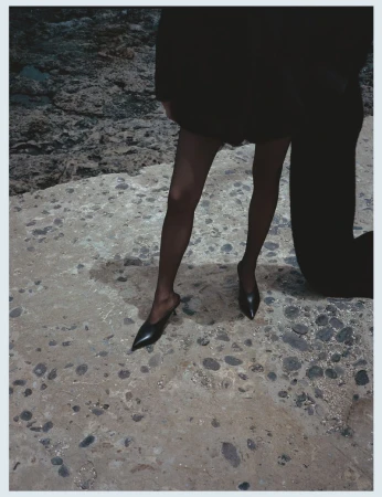 Fashion shot of a person in black tights and shoes, partially visible, standing on a textured stone surface with a rocky background.
