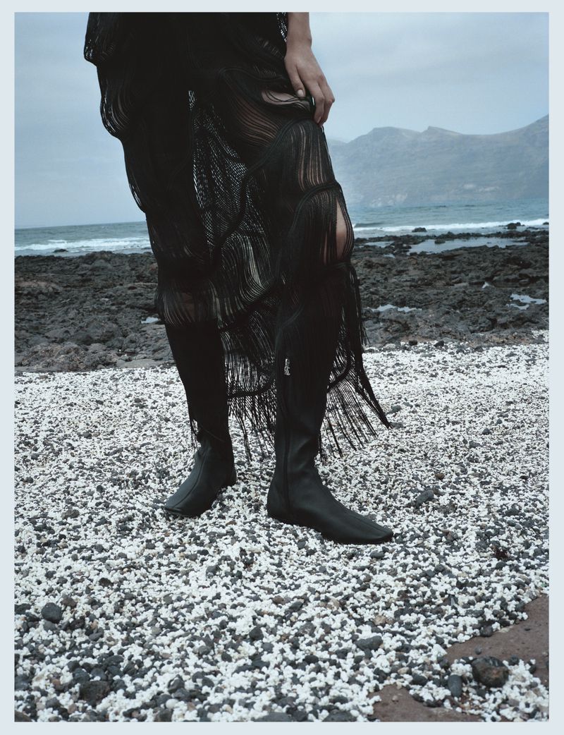Woman in a black dress and boots stands on a rocky beach with mountains and the ocean in the background, showcasing fashion and landscape.