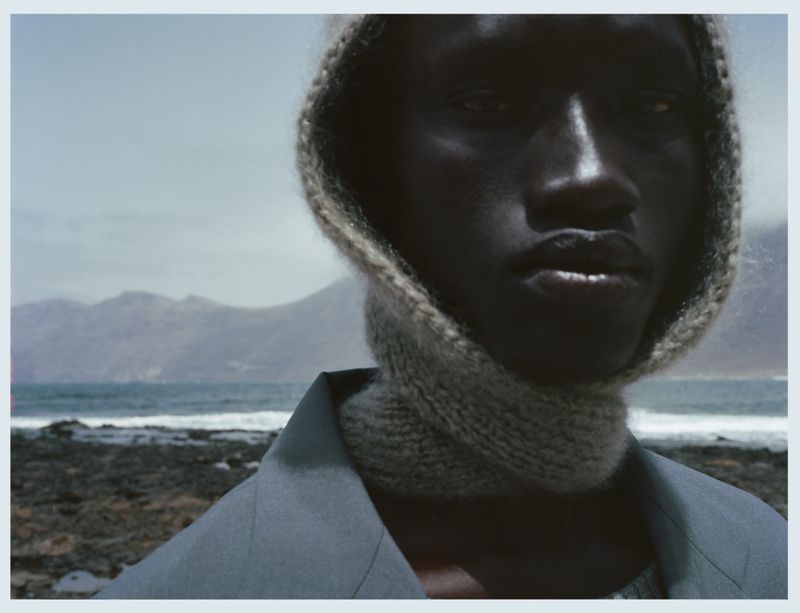 Portrait of dark-skinned person in knit hood and jacket, sea and mountain background, neutral hues and cinematic fashion style.