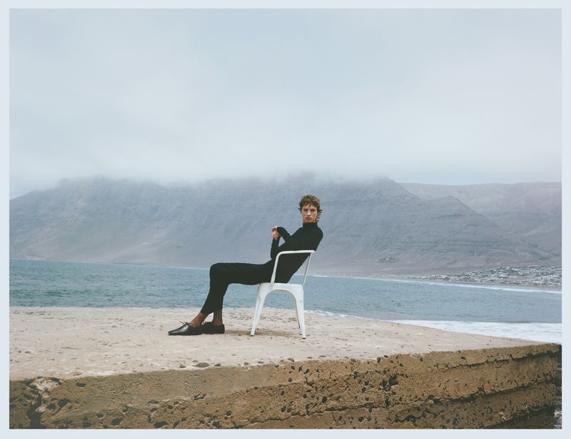Man in black turtleneck sitting on white chair on concrete pier with ocean and mountain backdrop on overcast day in Lanzarote, Spain.