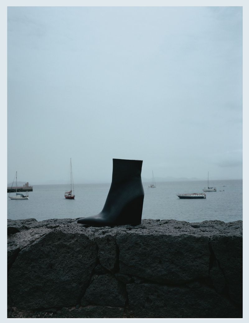 Black leather wedge boot displayed outdoors on a rock wall with sailboats on the water and a gray overcast sky in the background.