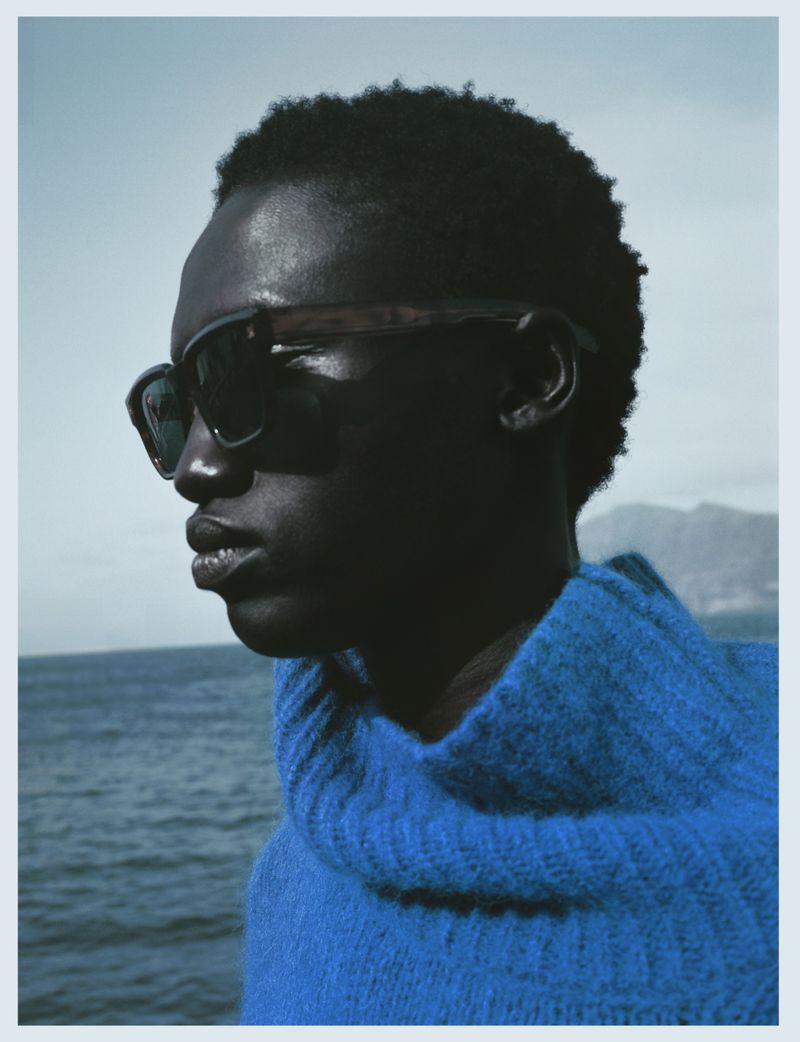 Close-up portrait of a Black male model with sunglasses and a blue sweater, posing against a sea and sky backdrop on a sunny day.