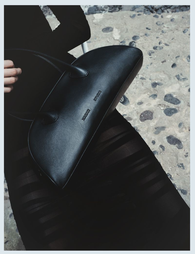 Fashion shot of a black leather handbag with a smooth finish held by a hand against a textured concrete background in a vertical format.
