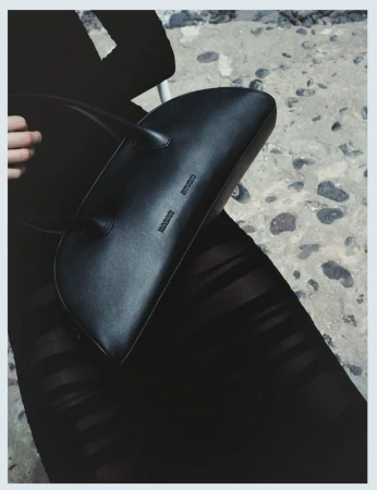 Fashion shot of a black leather handbag with a smooth finish held by a hand against a textured concrete background in a vertical format.