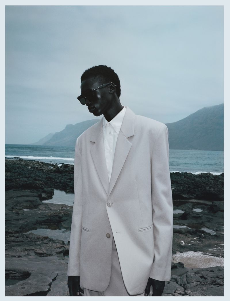 A solitary Black male model in a light grey suit poses on a rocky shore with mountains and ocean in the background under an overcast sky.