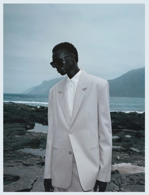 A solitary Black male model in a light grey suit poses on a rocky shore with mountains and ocean in the background under an overcast sky.