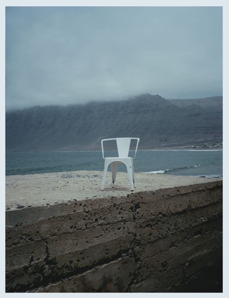 A minimalist landscape featuring a white chair on a concrete platform overlooking a sea and mountain range under a cloudy sky.