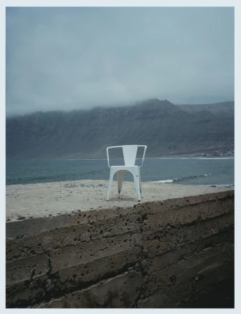 A minimalist landscape featuring a white chair on a concrete platform overlooking a sea and mountain range under a cloudy sky.