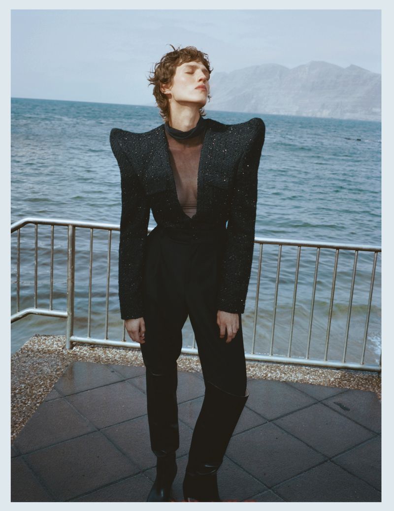 A person in a tailored dark jacket and trousers stands by a metal railing overlooking the water, with mountains in the distance.