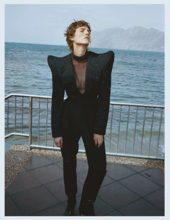 Model standing outdoors by the ocean in a structured black blazer with exaggerated shoulders and trousers.