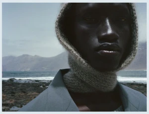 Close-up portrait of a black man wearing a knitted balaclava and grey blazer against an ocean and mountain landscape background.