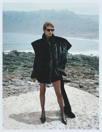 Model in black sunglasses and oversized black coat standing on a rocky coast by the ocean for Kazar Studio A/W 25 campaign.