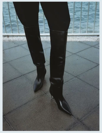 Close-up of black leather pointed-toe knee-high boots on a paved outdoor walkway overlooking water.