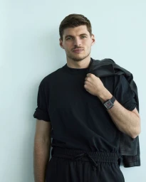 Max Verstappen wearing a black t-shirt and holding a black jacket against a plain wall for Tag Heuer