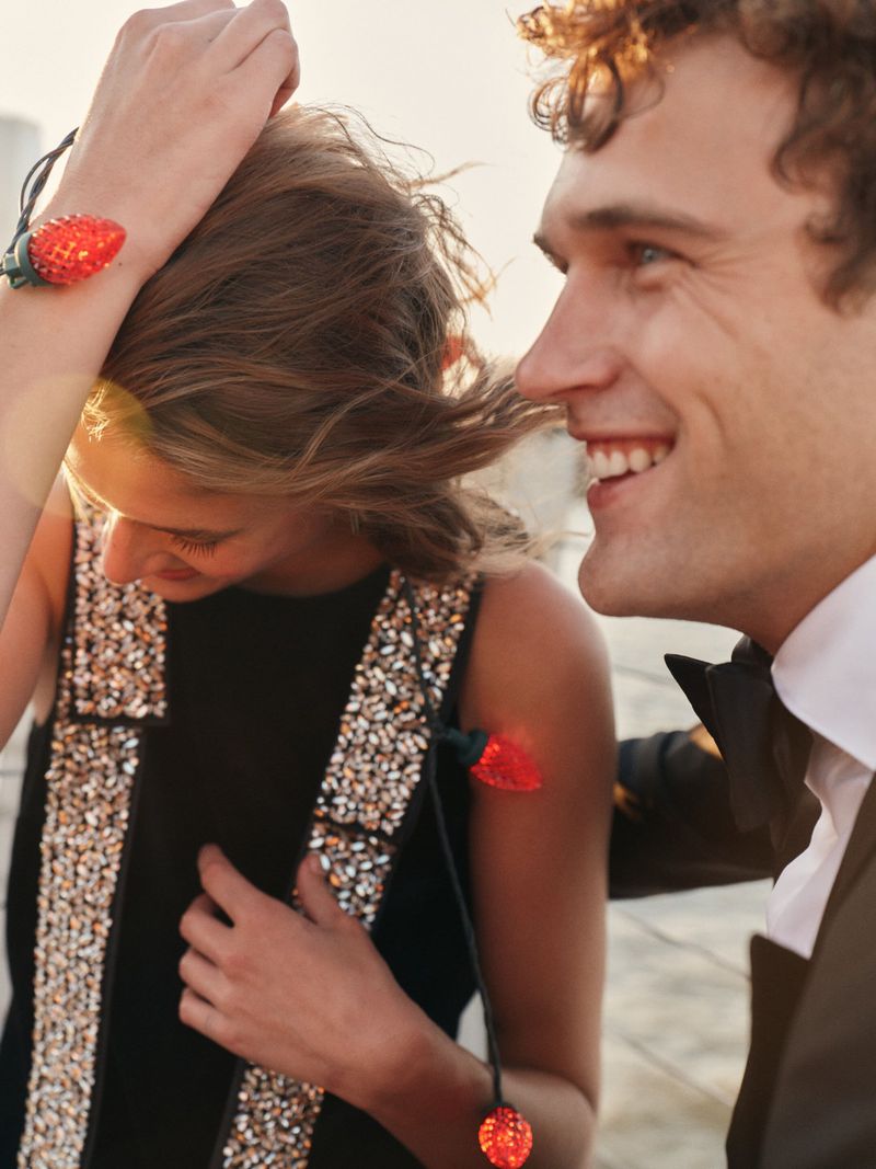 A smiling man and woman in formal wear stand together, with the woman wearing decorative strawberry lights on her dress and wrist.