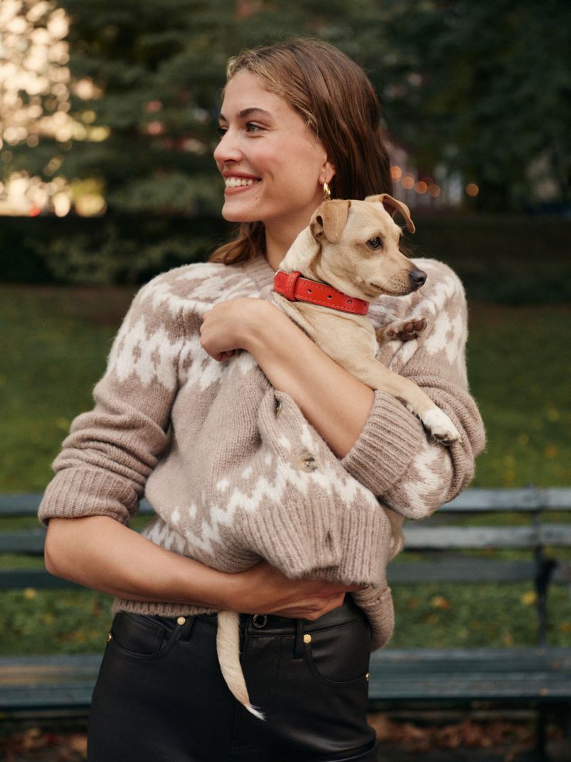 A woman in a fair isle sweater smiles while holding a small dog with a red collar outdoors in a park-like setting with trees and a bench.