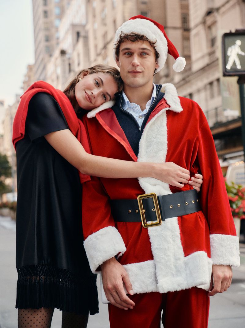 A young couple poses on a city street; the man wears a Santa suit, and the woman in black leans on him, wearing a red sweater.