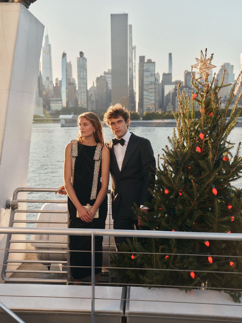 Woman in black dress with man in suit standing near Christmas tree with NYC skyline background. Formal fashion on boat deck.