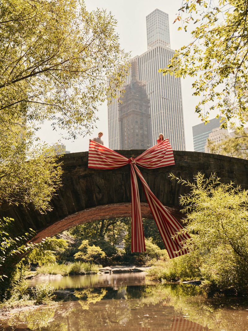 Scenic view of Central Park bridge adorned with red and white striped bow, with two people and skyscrapers in the background.