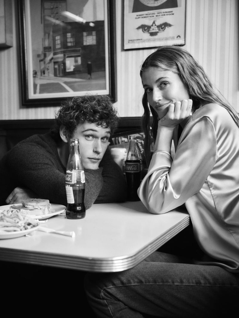 A black and white photograph captures a young couple in a vintage diner setting, seated at a table with food and Coca-Cola bottles.