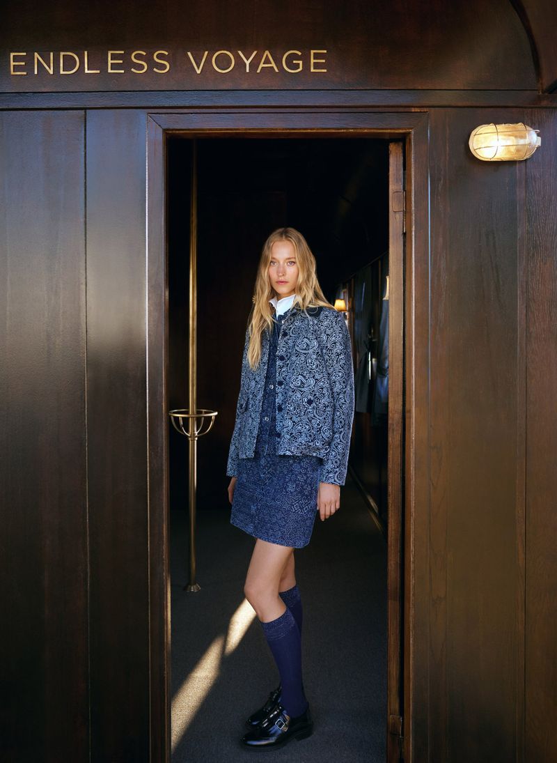 A young woman in a blue patterned jacket and matching skirt stands in a train doorway, looking at the camera in soft, diffused light.