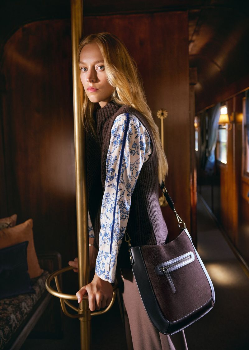 Indoor shot of a blonde adult woman in a vest and floral blouse holding a handrail with a purse on her shoulder looking at the camera.