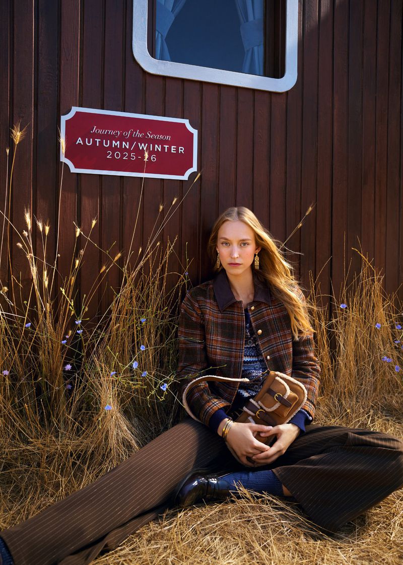 A fair-skinned woman in a plaid jacket and brown pants poses seated outdoors against a paneled wall in a fashion editorial shot.