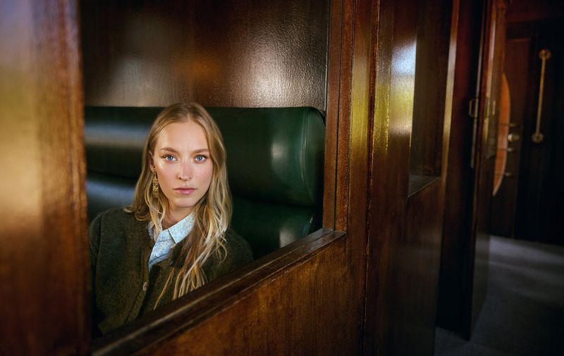 A young woman with long blonde hair sits inside of a vintage train car with a green leather seat and dark wood paneling looking forward.