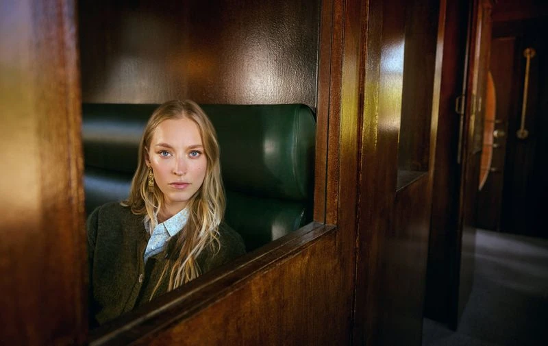 A young woman with long blonde hair sits inside of a vintage train car with a green leather seat and dark wood paneling looking forward.