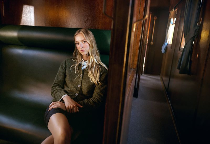 Young woman with long blonde hair sits inside a train car. She wears a sweater and skirt with a neutral expression, looking at the camera.