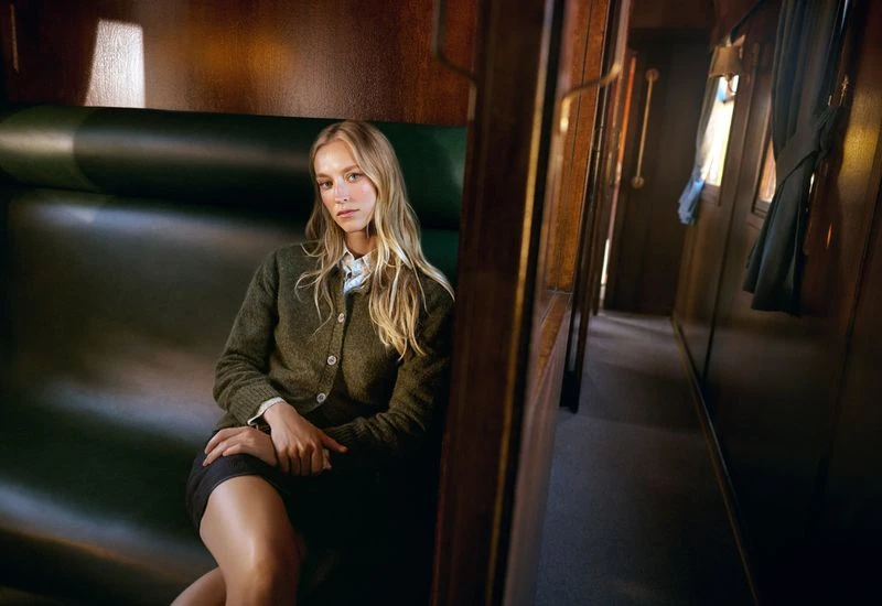 Young woman with long blonde hair sits inside a train car. She wears a sweater and skirt with a neutral expression, looking at the camera.