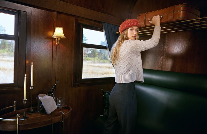 Woman in red beret places leather suitcase on luggage rack inside vintage train car with brown wood and green cushioned seats.