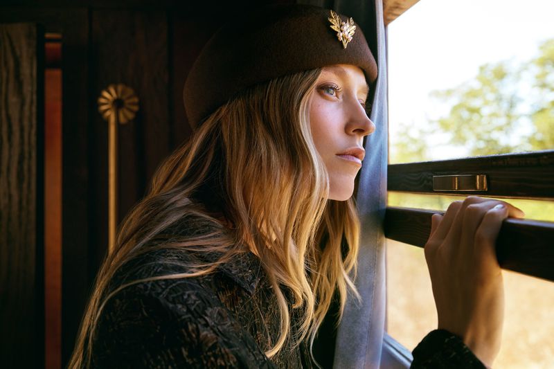 Adult woman with blonde hair and brown hat looking out of a train window on a sunny day.