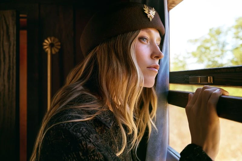 Adult woman with blonde hair and brown hat looking out of a train window on a sunny day.
