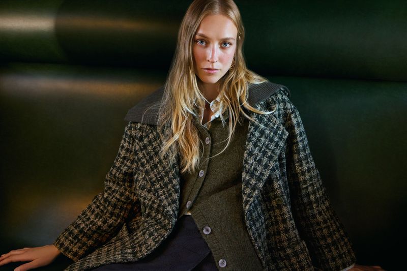 Portrait of a blonde woman in a patterned coat and cardigan, shot in a studio with a dark green background. Fashion editorial shot.