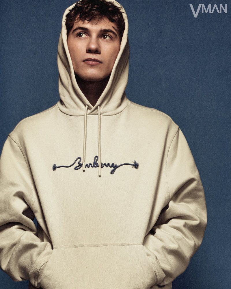 Portrait of a young man with curly brown hair, wearing a hooded sweatshirt with hands in pockets, looking up and to the left.