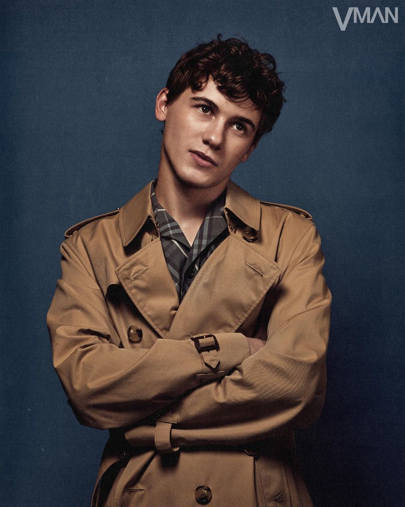 Portrait of a young adult male wearing a tan trench coat indoors against a blue backdrop. Fashion and style editorial photograph.