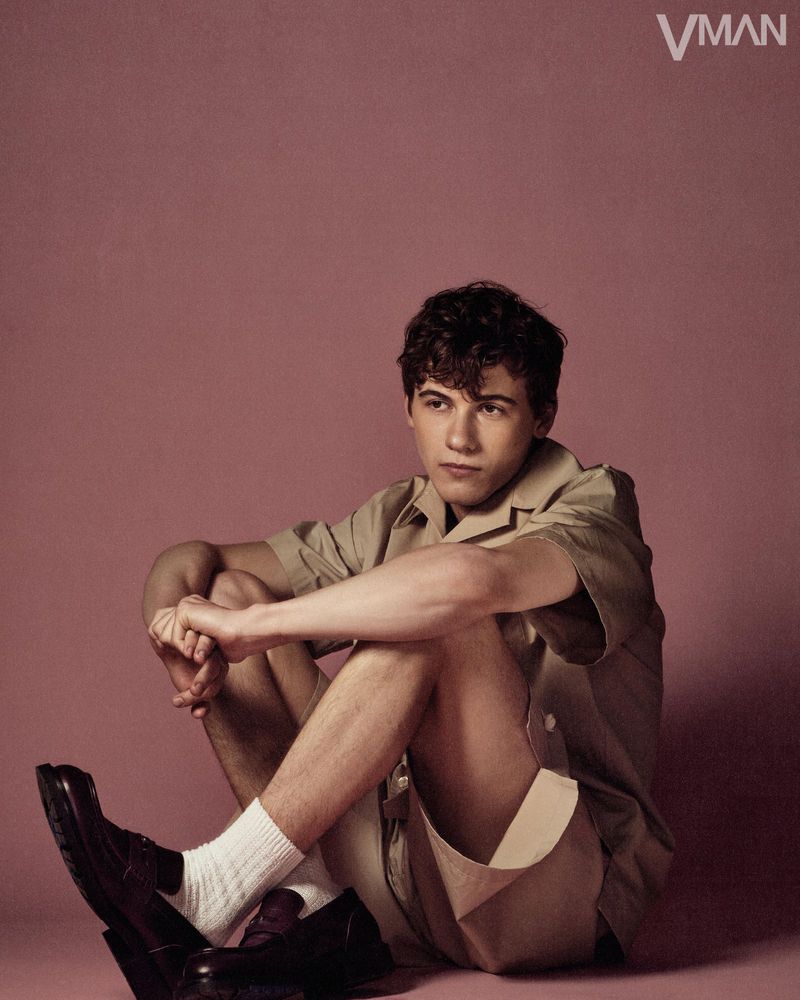 Sam Nivola seated on a solid muted background wearing a beige shirt, matching shorts, white socks, and dark loafers.