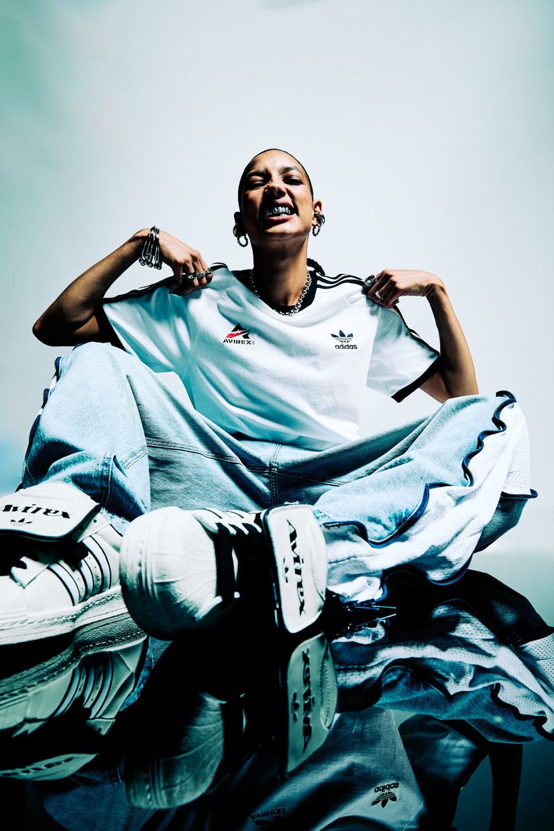 Stylish person with a shaved head poses with Adidas apparel: a t-shirt, loose jeans, and shoes displayed on a reflective surface.