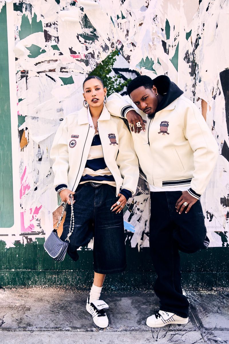 Two people, a man and a woman, pose in cream-colored varsity jackets in an outdoor setting with a torn poster wall background.