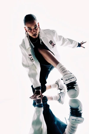 Stylish woman in white jacket poses with reflection wearing white Avirex sneakers. Fashion shot with white backdrop and high contrast lighting.
